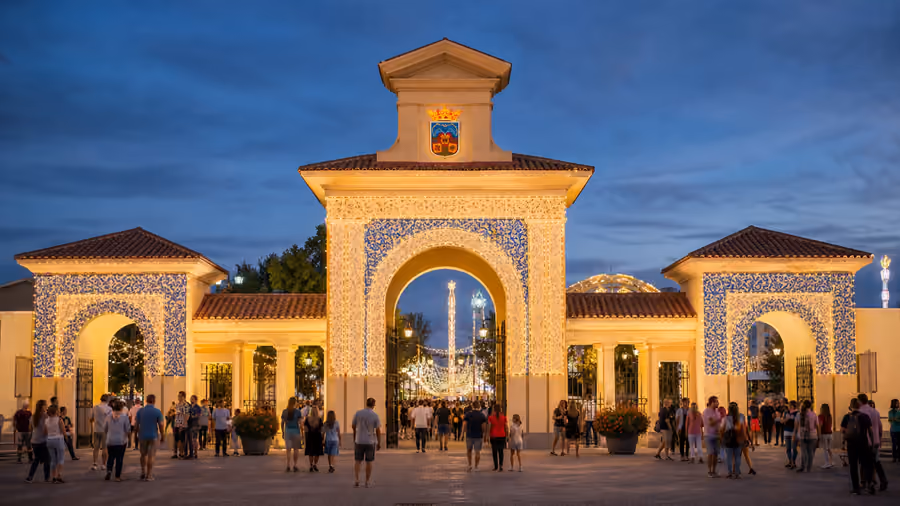 Recreación editorial de la puerta del recinto ferial iluminada al anochecer, con gente entrando bajo los arcos