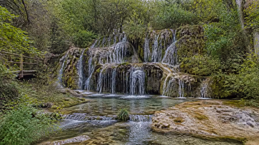 Recreación editorial de las cascadas del nacimiento del río Cuervo cayendo sobre roca cubierta de musgo, con una pasarela de madera a un lado