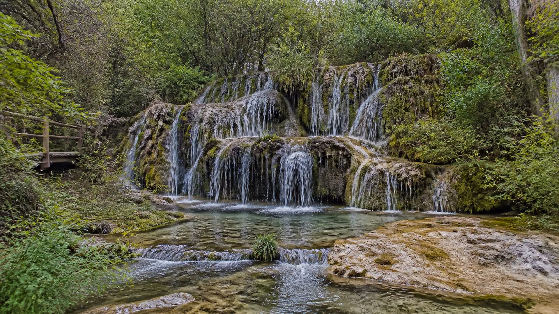 Recreación editorial de las cascadas del nacimiento del río Cuervo cayendo sobre roca cubierta de musgo, con una pasarela de madera a un lado