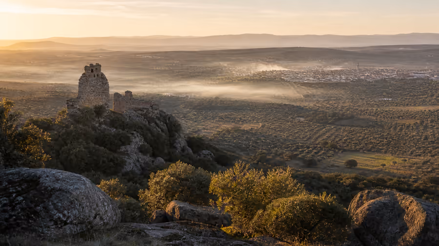 Recreación editorial de una torre en ruinas sobre un risco al amanecer, con niebla baja sobre los olivares