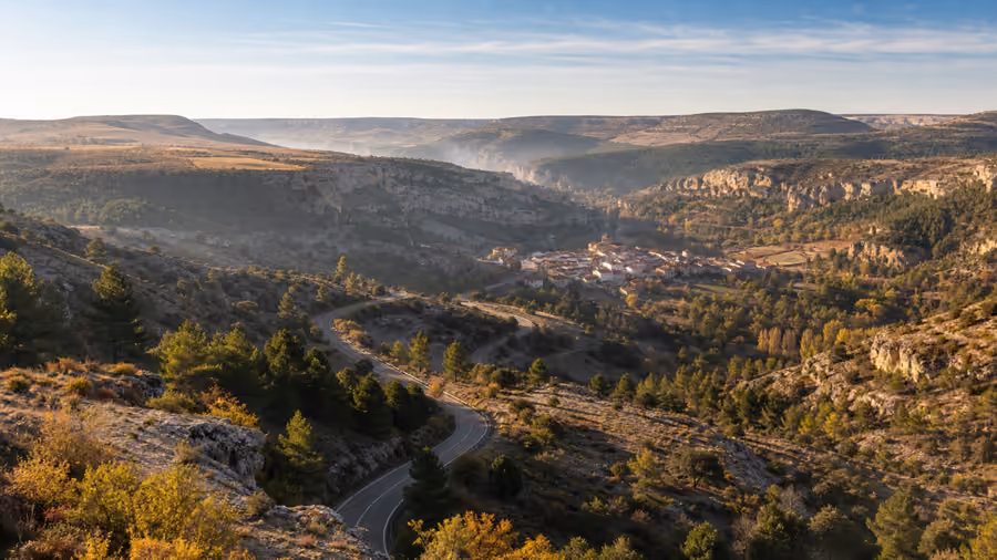 Recreación editorial de una carretera de curvas sobre un valle encajado, con un pueblo pequeño en el fondo