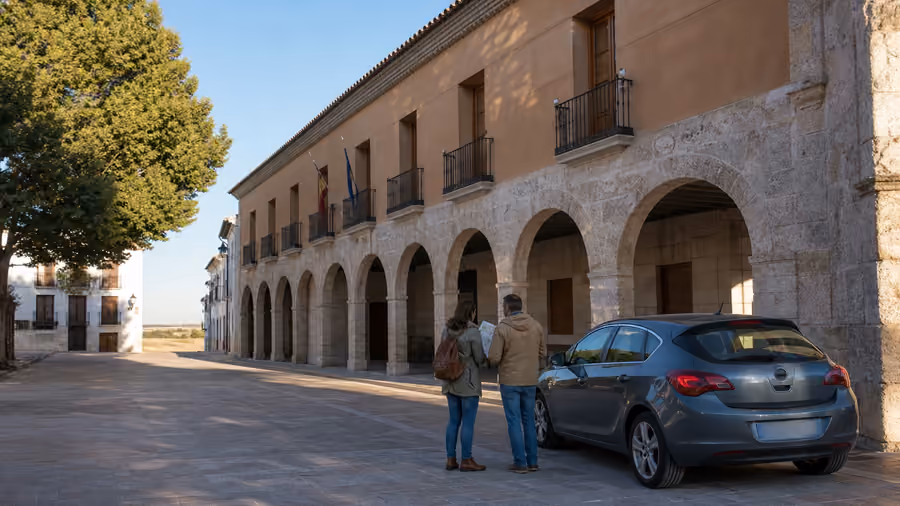 Recreación editorial de una pareja consultando un mapa junto a su coche, frente a un edificio con soportales de piedra