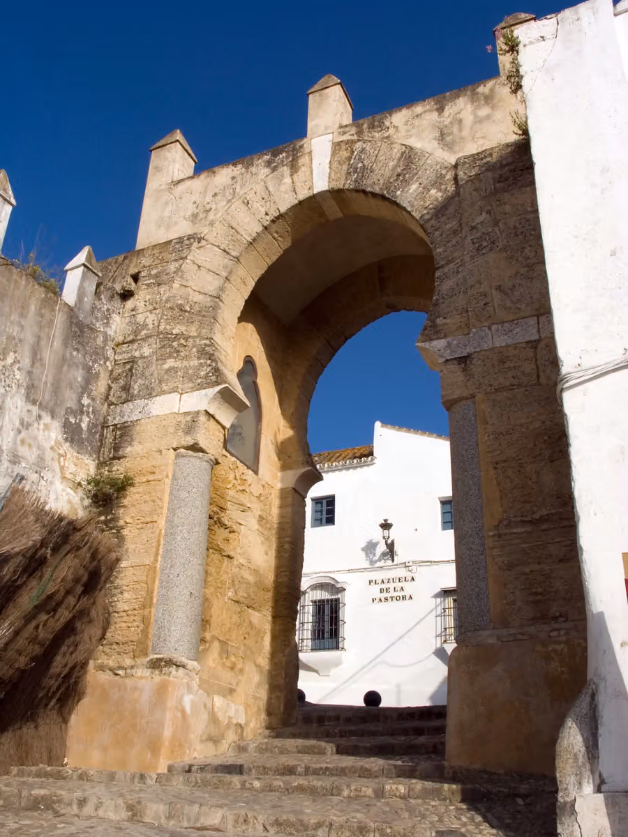 Arco de la Pastora de Medina Sidonia visto desde las escaleras, con muros de piedra y una fachada blanca al fondo